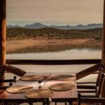 An image showing a scenic outdoor dining setup on a wooden deck overlooking a tranquil lake and distant mountains.