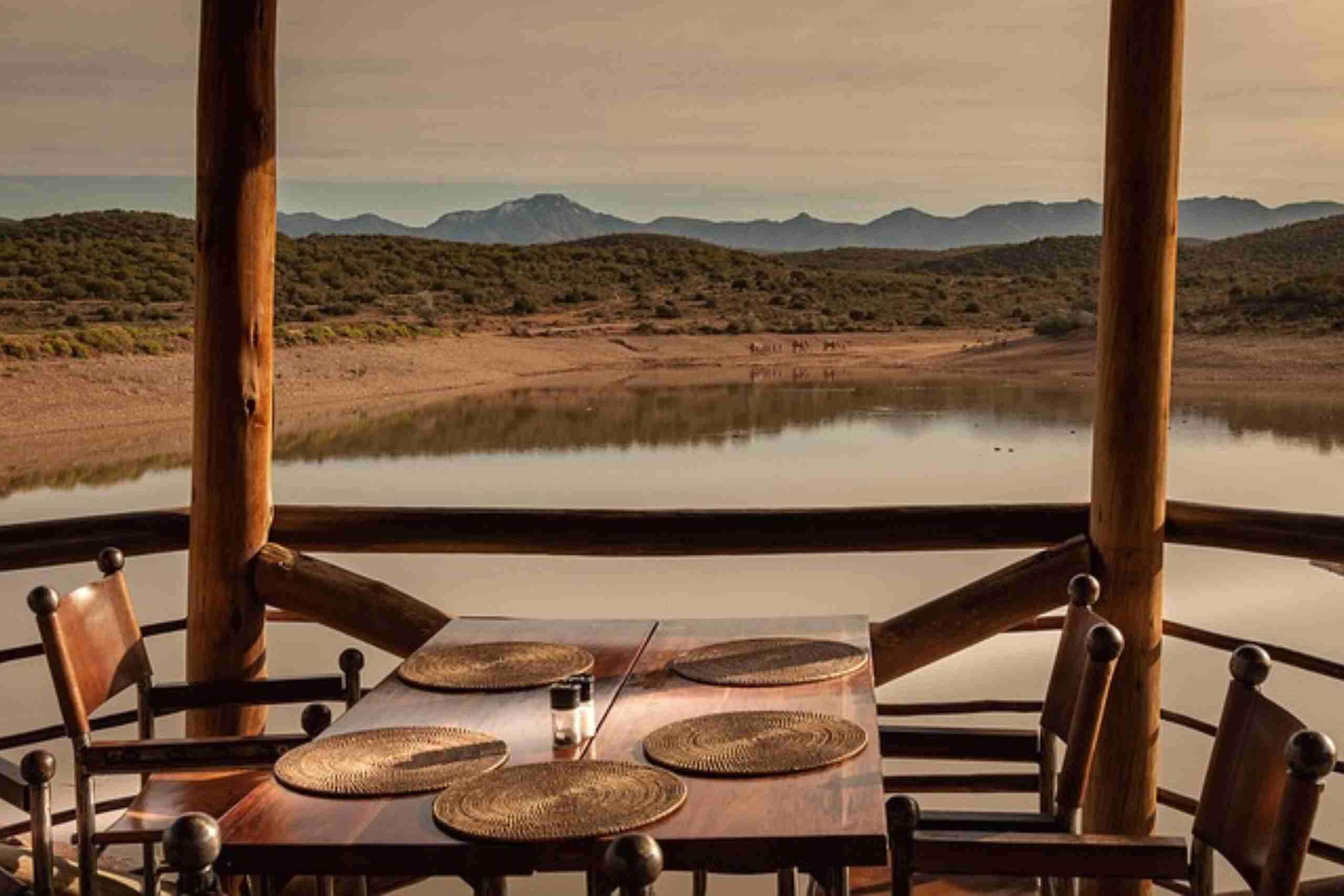 An image showing a scenic outdoor dining setup on a wooden deck overlooking a tranquil lake and distant mountains.